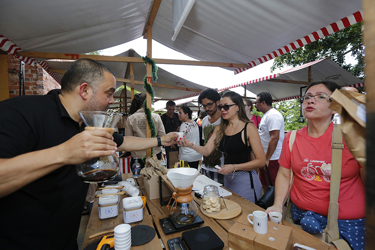 Barraca com degustação de café no Festival de Café. Homem de camisa preta oferencendo café a uma mulher, ao lado outro mulher de camisa vermelha bebendo café. 